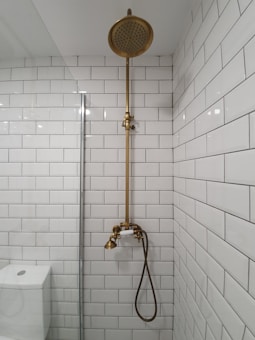 A bathroom with white subway tiles covering the walls, a vintage-style brass shower fixture that includes a showerhead at the top and a faucet below, and a modern toilet partially visible to the left. The overall look is clean and minimalistic.