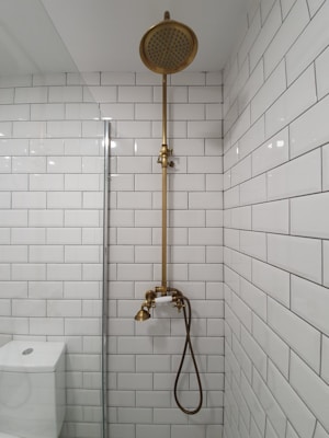A bathroom with white subway tiles covering the walls, a vintage-style brass shower fixture that includes a showerhead at the top and a faucet below, and a modern toilet partially visible to the left. The overall look is clean and minimalistic.