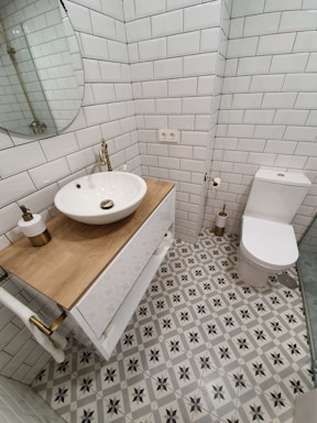 A small, modern bathroom featuring white subway tiles on the walls and a patterned tile floor with grey and black star-like designs. There is a wooden countertop with a round, white vessel sink and brass fixtures. A round mirror is mounted above the sink. The toilet is positioned next to the sink, with a toilet paper holder and a small, cylindrical brass trash can nearby. The overall design is clean and minimalist.