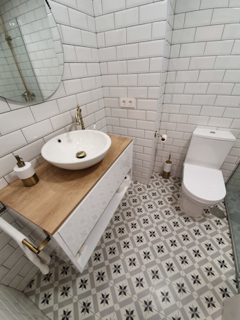 A small, modern bathroom featuring white subway tiles on the walls and a patterned tile floor with grey and black star-like designs. There is a wooden countertop with a round, white vessel sink and brass fixtures. A round mirror is mounted above the sink. The toilet is positioned next to the sink, with a toilet paper holder and a small, cylindrical brass trash can nearby. The overall design is clean and minimalist.