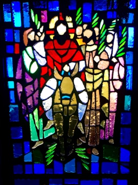 A stained glass artwork depicting a central figure in red and white robes surrounded by four other figures holding green branches. The background consists of geometric patterns in vibrant blue hues.