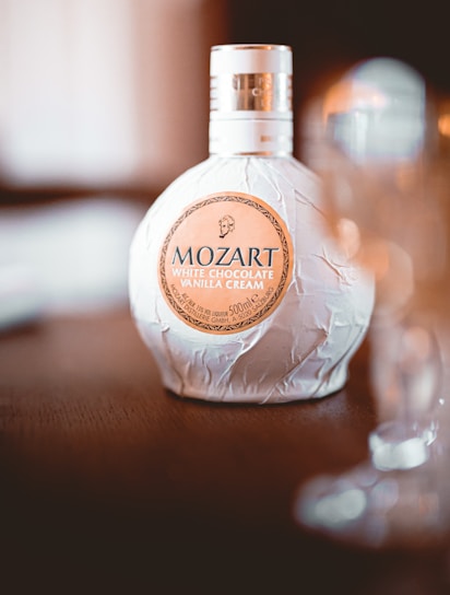A bottle of Mozart White Chocolate Vanilla Cream liqueur sits on a wooden surface with its decorative packaging. The background is softly blurred, adding elegance to the presentation.