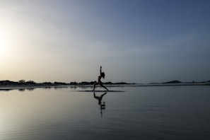 A person is practicing yoga in a warrior pose on the beach, reflected on the calm water. The sky is vast with a blend of light and darker shades, creating a serene and peaceful atmosphere. The horizon is lined with distant trees and a gentle hill on the right.