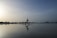 A person is practicing yoga in a warrior pose on the beach, reflected on the calm water. The sky is vast with a blend of light and darker shades, creating a serene and peaceful atmosphere. The horizon is lined with distant trees and a gentle hill on the right.