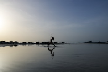 A person is practicing yoga in a warrior pose on the beach, reflected on the calm water. The sky is vast with a blend of light and darker shades, creating a serene and peaceful atmosphere. The horizon is lined with distant trees and a gentle hill on the right.