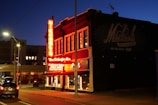 A brightly lit clothing store with a neon sign on the corner of a street at dusk. The store has a vintage appearance with signs advertising men's clothing items like Florsheim shoes, and Stetson hats. The warm glow of the neon lights contrasts with the blue evening sky and casts light onto the sidewalk and nearby parked cars.