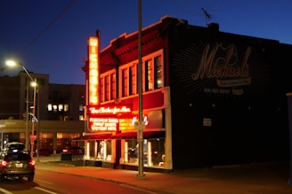 A brightly lit clothing store with a neon sign on the corner of a street at dusk. The store has a vintage appearance with signs advertising men's clothing items like Florsheim shoes, and Stetson hats. The warm glow of the neon lights contrasts with the blue evening sky and casts light onto the sidewalk and nearby parked cars.