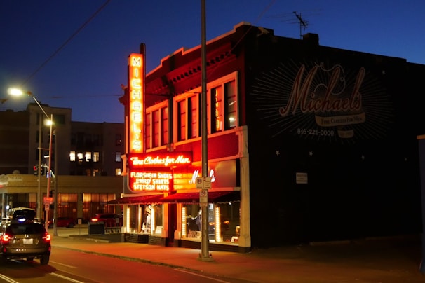 A brightly lit clothing store with a neon sign on the corner of a street at dusk. The store has a vintage appearance with signs advertising men's clothing items like Florsheim shoes, and Stetson hats. The warm glow of the neon lights contrasts with the blue evening sky and casts light onto the sidewalk and nearby parked cars.