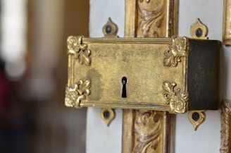 An ornate brass lock plate with intricate floral patterns and a keyhole is affixed to a decorative door. The metal has an aged, antique finish, highlighting its craftsmanship and vintage appeal.