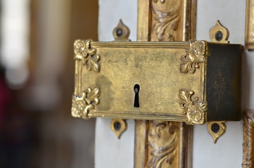 An ornate brass lock plate with intricate floral patterns and a keyhole is affixed to a decorative door. The metal has an aged, antique finish, highlighting its craftsmanship and vintage appeal.