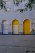 An exterior view of a building with distinct architectural features, including three archways. Two of the archways are sealed with bright yellow wooden panels, while the middle one has a white door. Text in capital letters is visible on the wall beside the archway on the left. Overhanging green tree branches create a natural frame along the top edge of the image.