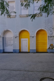 An exterior view of a building with distinct architectural features, including three archways. Two of the archways are sealed with bright yellow wooden panels, while the middle one has a white door. Text in capital letters is visible on the wall beside the archway on the left. Overhanging green tree branches create a natural frame along the top edge of the image.