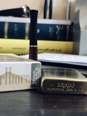A close-up shot of a pack of Marlboro cigarettes beside a brass Zippo lighter, with books in the background. A black and brown electronic device is also visible, possibly a vape or a pen.