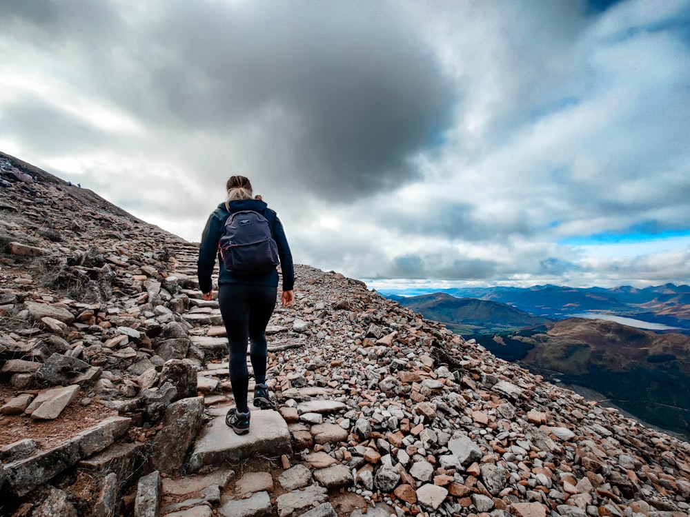 man in black jacket and black pants standing on rocky mountain under white clouds during daytime