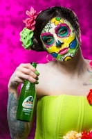 A person with elaborate and colorful face paint in the style of Dia de los Muertos, featuring floral and skull motifs. The individual is holding a green bottle of Jarritos grapefruit soda. They wear a bright green strapless top and have decorative flowers in their hair. The background is a vivid magenta color.