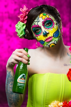A person with elaborate and colorful face paint in the style of Dia de los Muertos, featuring floral and skull motifs. The individual is holding a green bottle of Jarritos grapefruit soda. They wear a bright green strapless top and have decorative flowers in their hair. The background is a vivid magenta color.