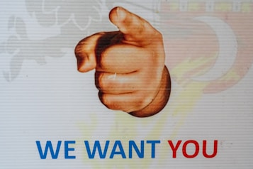 A poster with an image of a hand pointing forward. Below the hand, the text 'WE WANT YOU' is written, with 'WE WANT' in blue and 'YOU' in red.