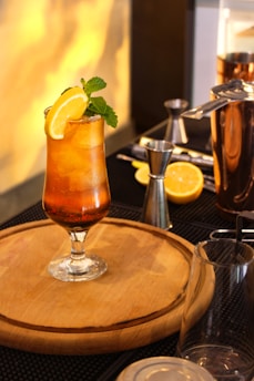 A refreshing cocktail sits on a wooden board, garnished with mint leaves and a lemon slice. Surrounding the drink are cocktail-making tools, including a jigger, shaker, and cut lemon slices. Warm lighting creates a cozy ambiance.