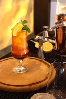 A refreshing cocktail sits on a wooden board, garnished with mint leaves and a lemon slice. Surrounding the drink are cocktail-making tools, including a jigger, shaker, and cut lemon slices. Warm lighting creates a cozy ambiance.