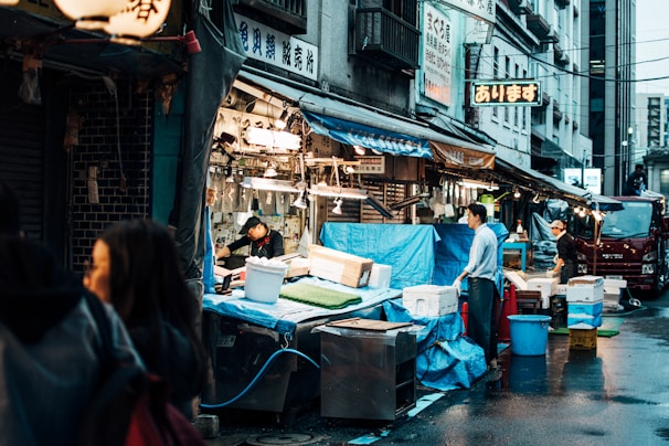 A bustling outdoor market scene with several vendors working at their stalls. The area is covered with blue tarps and contains various containers and boxes. Signs with Japanese characters are hanging above, and there is visible interaction among people, suggesting a lively atmosphere.
