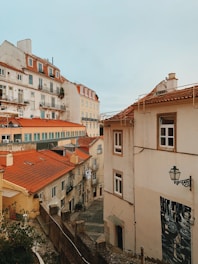 A view of a quaint European neighborhood with closely packed, multi-story residential buildings featuring red-tiled roofs. The architecture is classic, with white and beige walls, along with small balconies adorned with plants. A narrow cobblestone street winds through the buildings, and an antique street lamp is seen mounted on a wall.