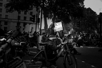 A black and white photograph of a public protest, featuring people gathered on the street. A sign is prominently displayed reading 'Racism is a Virus'. The crowd includes individuals wearing masks, emphasizing the seriousness and solidarity of the moment. Several bicycles can be seen, and the scene is set against an urban backdrop with buildings and trees.