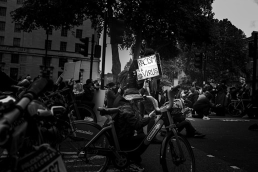A black and white photograph of a public protest, featuring people gathered on the street. A sign is prominently displayed reading 'Racism is a Virus'. The crowd includes individuals wearing masks, emphasizing the seriousness and solidarity of the moment. Several bicycles can be seen, and the scene is set against an urban backdrop with buildings and trees.