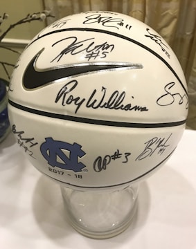 A white basketball decorated with numerous signatures and a large Nike logo. It features the logo and letters of the University of North Carolina, dated 2017-18. The ball is placed on a clear stand and is set against a neutral background.