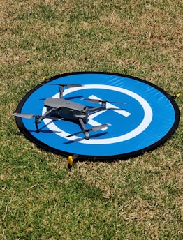 A small consumer drone is positioned on a circular blue landing pad marked with white design. The landing pad is placed on the grass, indicating an outdoor setting under clear daylight.
