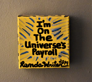 A small yellow canvas displays bold black text that reads 'I'm on the Universe's Payroll' with the artist's signature and date at the bottom. There are blue and gray radial lines emanating from the text, creating a dynamic visual effect.