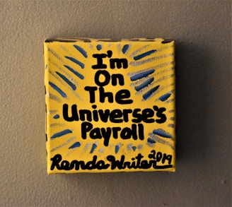 A small yellow canvas displays bold black text that reads 'I'm on the Universe's Payroll' with the artist's signature and date at the bottom. There are blue and gray radial lines emanating from the text, creating a dynamic visual effect.