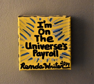 A small yellow canvas displays bold black text that reads 'I'm on the Universe's Payroll' with the artist's signature and date at the bottom. There are blue and gray radial lines emanating from the text, creating a dynamic visual effect.