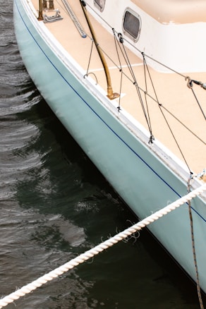 A close-up view of a sailboat's deck, featuring light blue and beige colors. The deck has metallic railings and black ropes attached. The boat is positioned on dark, rippling water, secured by a thick white rope.