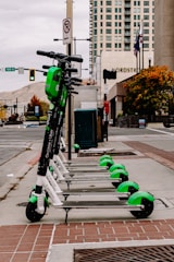 A row of electric scooters is neatly lined up on the sidewalk in an urban setting. In the background, a tall building and various street signs and traffic lights are visible. The scooters are predominantly white and green, and the street appears to be relatively quiet.
