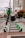 A row of electric scooters is neatly lined up on the sidewalk in an urban setting. In the background, a tall building and various street signs and traffic lights are visible. The scooters are predominantly white and green, and the street appears to be relatively quiet.