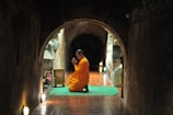 A person in an orange robe kneels in prayer on a green mat inside a dimly lit, narrow tunnel with brick walls. Soft lighting is visible on the sides, creating a peaceful and reflective ambiance, while an altar with flowers is placed to the side.