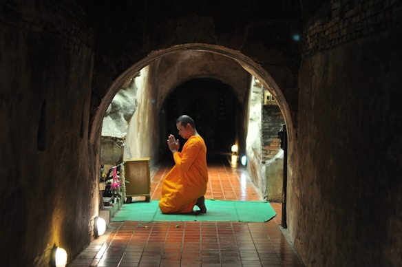 A person in an orange robe kneels in prayer on a green mat inside a dimly lit, narrow tunnel with brick walls. Soft lighting is visible on the sides, creating a peaceful and reflective ambiance, while an altar with flowers is placed to the side.