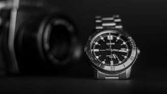 A black and silver wristwatch with a clear, detailed dial is positioned prominently in the foreground. The metal bracelet and the watch face display precise time indicators and the brand name. In the blurred background, there is a camera, suggesting a theme of precision and craftsmanship.