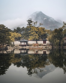 A traditional Korean pavilion is situated near a serene pond with trees surrounding it. The scene is reflected in the calm water. Mist enshrouds a mountain peak in the background, enhancing the tranquil ambiance.