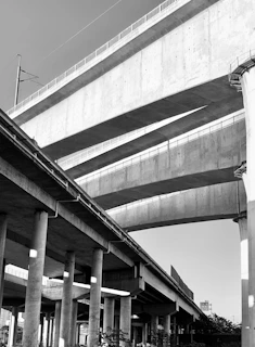 A black and white image of large, towering concrete bridges with multiple layers, supported by numerous pillars. The bridges rise high into the sky, intersecting one another, with architectural lines and shadows creating a dramatic visual effect.
