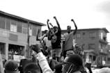 A group of people are standing on a structure in an urban setting, raising their fists in the air, suggesting a protest or demonstration. The crowd appears engaged and determined, with some individuals taking photos or videos. The background features residential buildings.