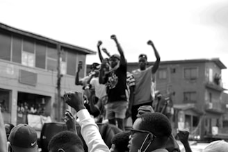 A group of people are standing on a structure in an urban setting, raising their fists in the air, suggesting a protest or demonstration. The crowd appears engaged and determined, with some individuals taking photos or videos. The background features residential buildings.