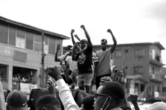 A group of people are standing on a structure in an urban setting, raising their fists in the air, suggesting a protest or demonstration. The crowd appears engaged and determined, with some individuals taking photos or videos. The background features residential buildings.