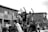 A group of people are standing on a structure in an urban setting, raising their fists in the air, suggesting a protest or demonstration. The crowd appears engaged and determined, with some individuals taking photos or videos. The background features residential buildings.