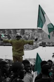A person stands elevated above a crowd, arms outstretched, holding a large flag resembling the Nigerian flag. The crowd appears dense, with people looking in various directions, and there is a banner in the background with text. The image predominantly uses black and white with selective coloring for the flag.