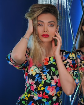 A person with long blonde hair, vibrant red lipstick, and expressive eyebrows is posing confidently. They are wearing a colorful floral dress, featuring a variety of bright colors. They appear to be in a space with reflective surfaces and deep blue backgrounds, adding a modern and stylish touch.
