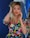 A person with long blonde hair, vibrant red lipstick, and expressive eyebrows is posing confidently. They are wearing a colorful floral dress, featuring a variety of bright colors. They appear to be in a space with reflective surfaces and deep blue backgrounds, adding a modern and stylish touch.