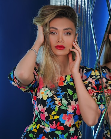 A person with long blonde hair, vibrant red lipstick, and expressive eyebrows is posing confidently. They are wearing a colorful floral dress, featuring a variety of bright colors. They appear to be in a space with reflective surfaces and deep blue backgrounds, adding a modern and stylish touch.