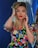 A person with long blonde hair, vibrant red lipstick, and expressive eyebrows is posing confidently. They are wearing a colorful floral dress, featuring a variety of bright colors. They appear to be in a space with reflective surfaces and deep blue backgrounds, adding a modern and stylish touch.