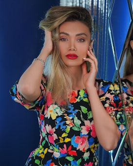 A person with long blonde hair, vibrant red lipstick, and expressive eyebrows is posing confidently. They are wearing a colorful floral dress, featuring a variety of bright colors. They appear to be in a space with reflective surfaces and deep blue backgrounds, adding a modern and stylish touch.